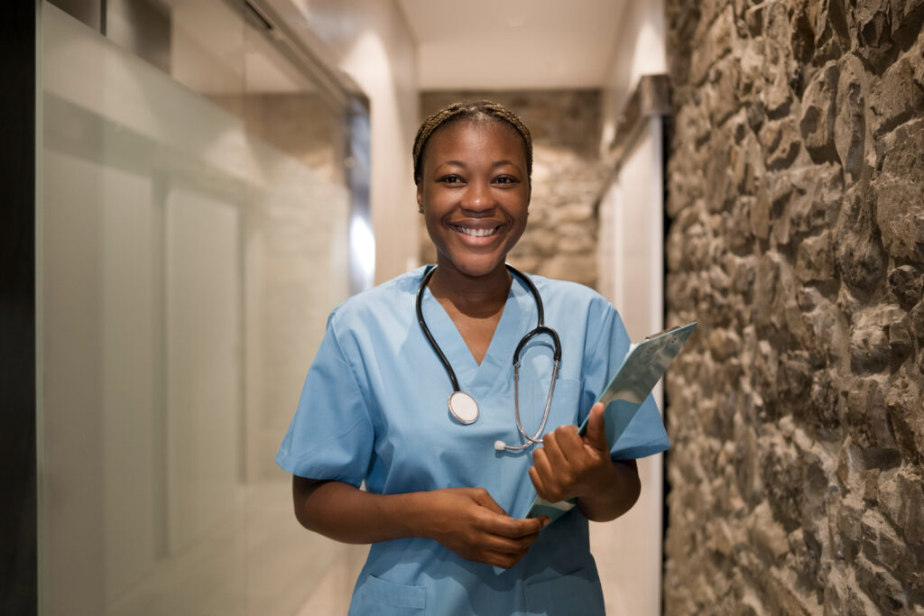 mulher negra sorrindo vestindo avental de hospital segurando uma prancheta e usando um estetoscópio. Ao fundo corredor de um hospital. imperva waf